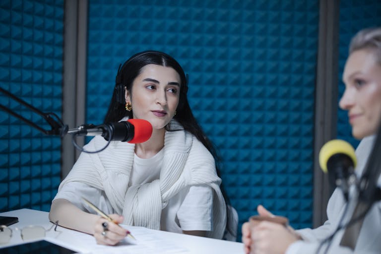Two women engaged in a podcast recording with professional microphones, soundproof studio.